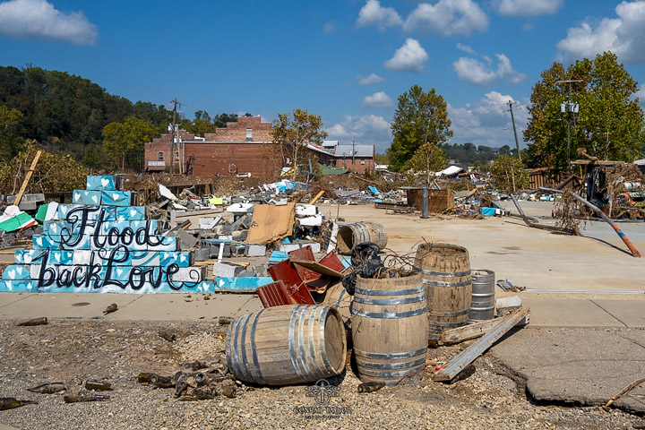 Photo by Conrad Taylor Photography showing the aftermath of Hurricane Helene in the River Arts District, Asheville. Wooden barrels, debris, and a mural with the message 'Flood Back Love' amidst the destruction.