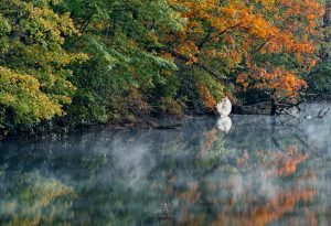 A quiet river in Highlands, NC, with mist rising off the water and autumn leaves reflecting in the river. A white canoe rests peacefully under the trees. Photo by: Conrad Taylor Photography.
