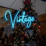 A glowing neon sign displaying the word 'Vintage,' illuminated in bright blue, hanging in a shop window with a backdrop of holiday decorations, including a Christmas tree and snowflake ornaments.