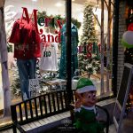 A holiday window display featuring mannequins dressed in seasonal outfits, framed by festive decor like Christmas trees, an elf figure, and a bench in front, with 'Merry and Bright' text on the glass.