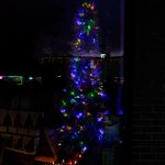 A Christmas tree adorned with vibrant multicolored lights, displayed in a darkened shop window with faint reflections of the surrounding town.