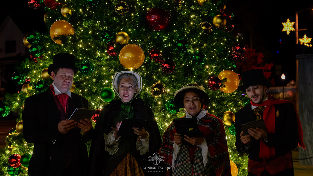 A group of carolers dressed in traditional Victorian attire singing in front of a brightly decorated Christmas tree, with gold and red ornaments glowing under the holiday lights.
